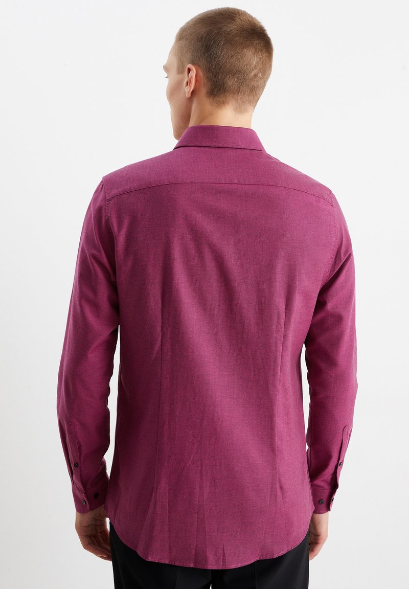 Man die met zijn rug naar de camera staat, gekleed in een donkerroze, lange mouw, knoopshirt en zwarte broek, tegen een eenvoudige witte achtergrond.