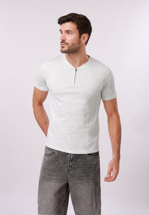 Muž s bradou, oblečený v svetlošedej košeli s krátkym rukávom a predným zipsom, s vybledlými čiernymi džínsami, pozerajúci sa doľava na jednoduchom pozadí.