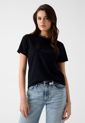 Jeune femme aux cheveux bruns portant un t-shirt Guess noir et un jean bleu clair, debout devant un fond blanc uni.