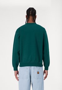 Green cotton sweatshirt with a crew neckline, ribbed cuffs, and hem. Worn with light blue denim pants featuring patch pockets.