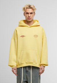 Žlutá bavlněná mikina s kapsou na přední straně, s vyšitým textem v hnědé barvě. Oversized střih a žebrované manžety. Nosená s volnými džínami.