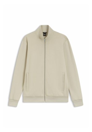 Beige vetoketjullinen takki, jossa pitkät hihat, pystykaulus, kaksi sivutaskua sekä resorit hihansuissa ja helmassa, aseteltuna tasaisesti valkoiselle taustalle.