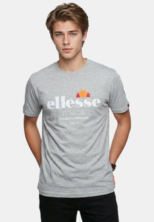 Jeune homme aux cheveux châtain clair portant un t-shirt gris avec le logo ellesse, debout les mains derrière le dos sur un fond blanc.