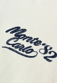 Texte brodé "Monte Carlo" en fil bleu marine sur un fond en tissu crème clair, avec un design en cursive et des points texturés.