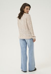 Chemise beige clair à manches longues avec un motif marbré blanc, associée à un jean évasé bleu clair. La chemise a des manches froncées et une coupe décontractée.