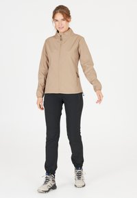 Beige softshell jack met een hoge kraag, een ritssluiting aan de voorkant en zijzakken, gecombineerd met zwarte tapered broek en lichte wandelschoenen.