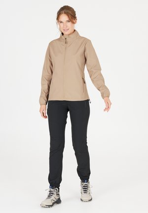 Beige mjukskaljacka med hög krage, framdragkedja och sidofickor, parat med svarta avsmalnande byxor och lätta vandringskängor.