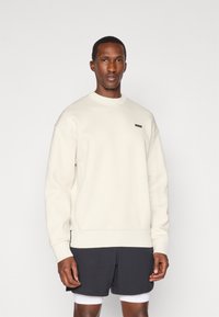 Sweatshirt crème avec un col et des poignets côtelés, arborant une petite étiquette noire. Associé à un short sombre et un sous-vêtement blanc.