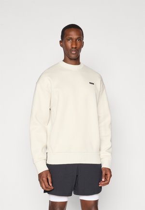 Sudadera crema con cuello y puños de canalé, con una pequeña etiqueta negra. Combinada con pantalones cortos oscuros y una camiseta blanca debajo.