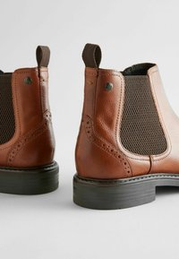 Botas de tobillo de cuero marrón con paneles laterales elásticos, un diseño texturizado y perforaciones decorativas, descansando sobre una suela gruesa de goma negra.