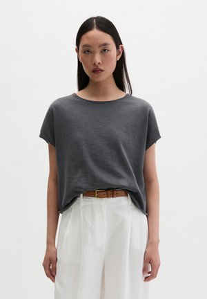 Woman with long straight black hair wearing a dark gray short-sleeve top tucked into high-waisted white pants with a brown belt.