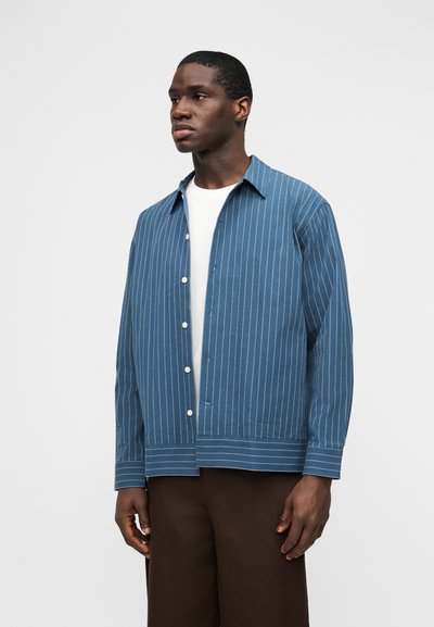 Άνδρας που φοράει μπλε πουκάμισο με λευκές ρίγες πάνω από λευκό πουκάμισο και καφέ παντελόνια, στέκεται μπροστά σε ένα απλό ανοιχτό γκρι φόντο.