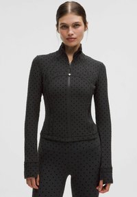 Chaqueta negra con cremallera fabricada en tejido elástico, presenta un patrón de puntos, diseño ajustado, cuello alto y mangas largas con orificios para los pulgares.