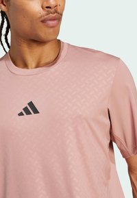 Svijetlo ružičasta atletska majica izrađena od prozračnog materijala, s suptilnim reljefnim uzorkom i crnim Adidas logom na lijevoj strani prsnog koša.