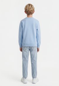 Blond kind draagt een lichtblauwe sweatshirt, lichtblauwe spijkerbroek en witte sneakers, staand met de rug naar de camera op een witte achtergrond.