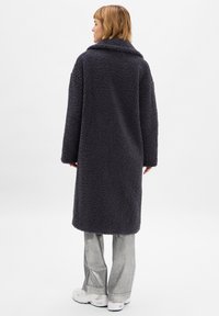 Donkergrijs, fluffy, oversized jas van textuurstof met lange mouwen en een brede kraag, gecombineerd met metallic grijze uitlopende broek.