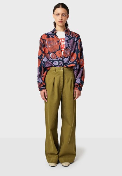 Žena stojaca, oblečená v olivových širokých nohaviciach, bielej grafickej tričku a voľnej košeli s kvetinovým vzorom v námornej, červenej a fialovej farbe.