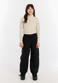 Lichtbeige fleece top met halve rits en hoge kraag; gecombineerd met zwarte, loszittende sneeuwbroek en zwarte veterschoenen.