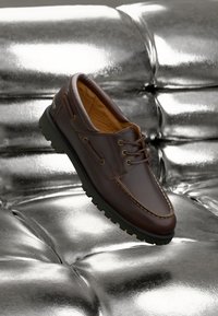 Shoe casual lace-up em couro castanho escuro com sola preta robusta, repousando sobre uma superfície prateada brilhante e estofada.