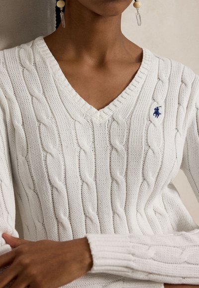 Persona valkā baltu pinumu V veida džemperi ar mazu tumši zilu izšūtu logotipu uz krūtīm un karājošām auskaru pāri.