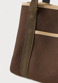 Καφέ τσάντα tote από τσόχα με μπεζ δερμάτινες λεπτομέρειες και πλατιές υφασμάτινες ελαιόχρωμες λουρίδες σε λευκό φόντο.