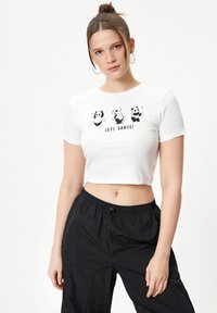 Camiseta corta blanca con textura acanalada, que presenta tres pandas negros y el texto "¡A BAILAR!" en negro. Combinada con pantalones negros de corte holgado.
