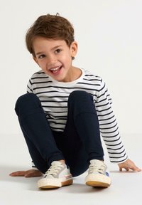 Dreng sidder på gulvet iført en hvid og marineblå stribet langærmet skjorte, marineblå bukser og beige sneakers med blå accenter.