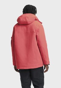 Asmuo su trumpais dreadsiais, vilkintis koralinę kapučiną ir juodus krovinius, atsisukęs nugarą priešais vientisą šviesų foną.