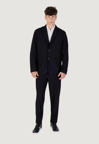 Giovane uomo con capelli ricci che indossa un abito nero, camicia bianca e sneaker nere, in piedi davanti a uno sfondo chiaro e semplice.