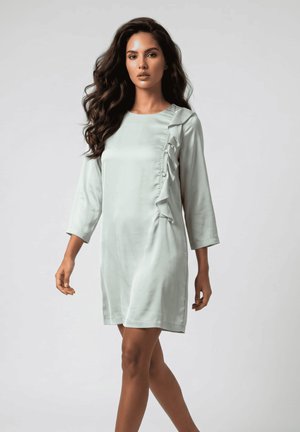 Mujer con cabello largo y ondulado que lleva un vestido gris claro hasta la rodilla con mangas tres cuartos y detalle de volantes en el lado derecho delantero.
