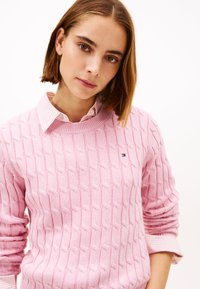 Pinkfarbener Pullover mit Zopfmuster, Rundhalsausschnitt und gerippten Bündchen, getragen über einem rosa-weiß gestreiften Hemd mit Kragen. Mit kleinem Logo.