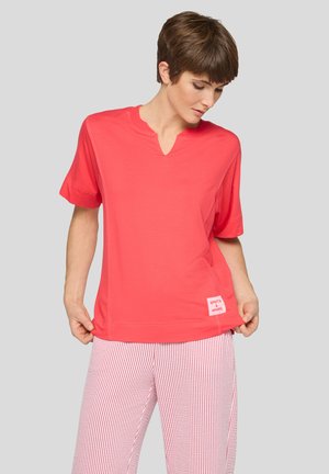 Person trägt ein korallenfarbenes kurzärmeliges Shirt mit Schlitz am Ausschnitt und rot-weiß gestreifte Hose, schaut nach unten und hält den Saum des Shirts.