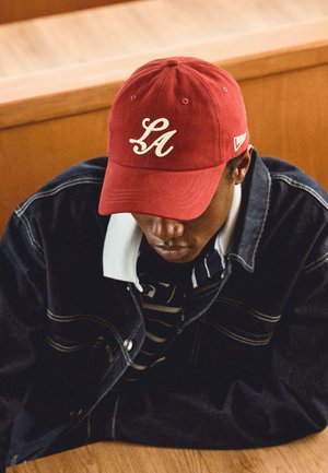 Mladi moški, ki nosi rdečo kapo z vezenim napisom "LA", oblečen v temno jeans jakno čez črtasto srajco, sedi za leseno mizo.