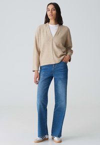 Maglione beige a maglia con scollatura a V, maniche lunghe e una vestibilità rilassata abbinato a jeans blue a gamba dritta e sneakers bianche con strisce nere.
