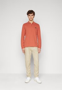 Polo a maniche lunghe in ruggine con colletto a contrasto, abbinato a pantaloni beige chiaro e sneaker bianche. Design semplice, vestibilità casual.