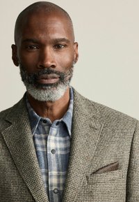 Sacou pentru bărbați dintr-un material maro texturat, peste o cămașă cu carouri albastre. Detalii remarcabile includ un batistă de buzunar cu un model geometric.