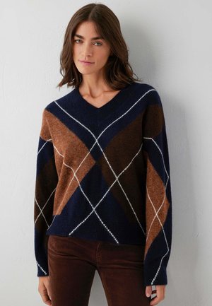 Frau trägt einen marineblauen und braunen Argyle-Muster V-Ausschnitt-Pullover und dunkelbraune Hose, steht vor einer weißen Wand.