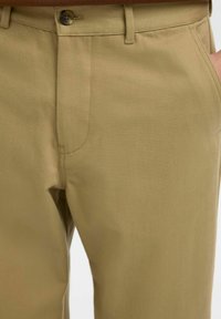 Gros plan sur un pantalon beige montrant la ceinture avec un bouton, des passants pour ceinture, une couture avant, et une main partiellement dans la poche droite.