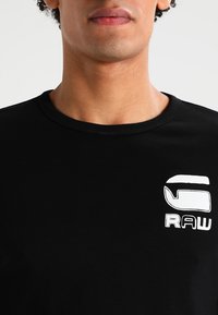 Close-up van een persoon die een zwart T-shirt draagt met een wit "G RAW" logo op de linkerborst, tegen een effen lichte achtergrond.