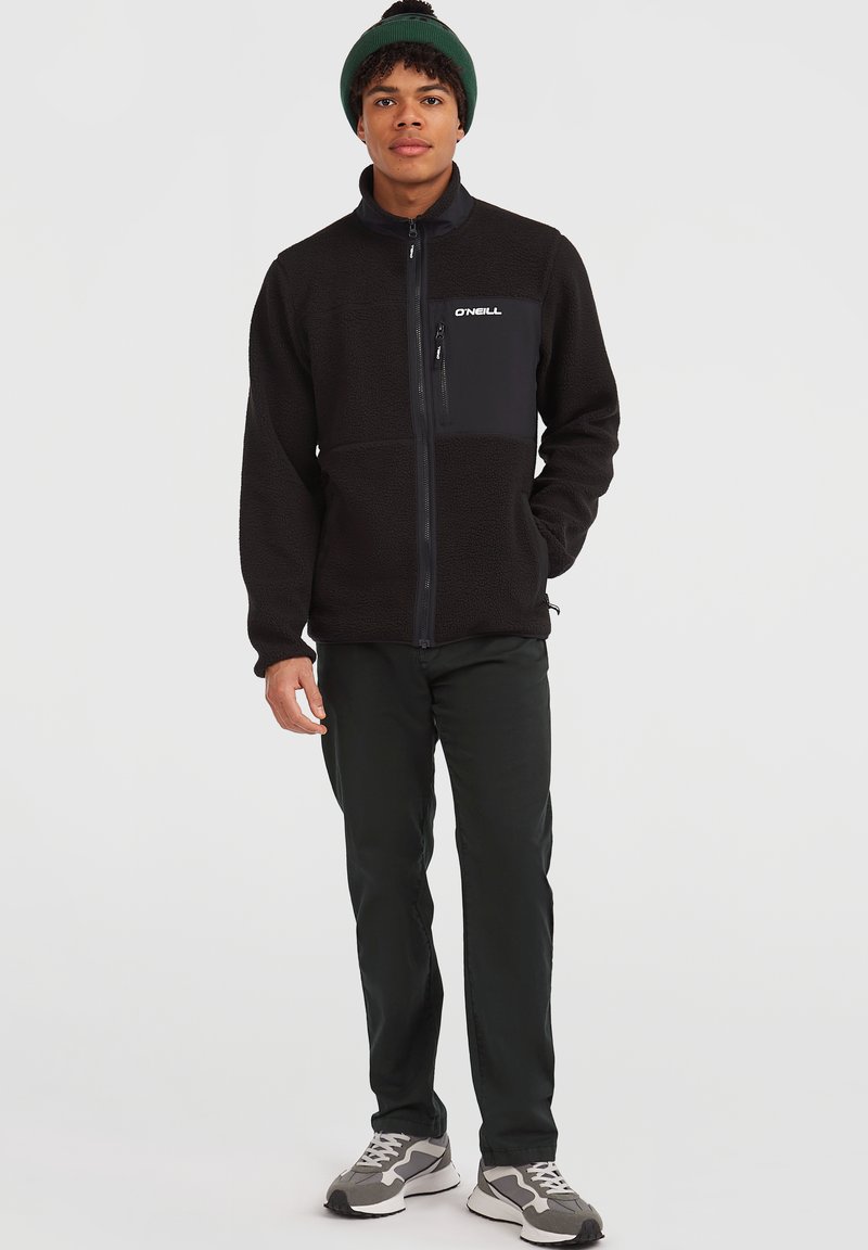 Giovane uomo con un berretto verde, giacca in pile nera con zip e logo sul taschino, pantaloni scuri e sneakers grigie, in piedi davanti a uno sfondo neutro.
