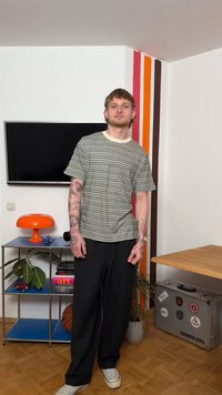 Tânăr cu tatuaje poartă o cămașă cu modele și pantaloni negri, stând într-o cameră cu televizor, lampă portocalie, plante și colț de perete cu dungi colorate.