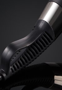 Gros plan sur une poignée de sèche-cheveux noire et chromée avec des fentes de ventilation sur un fond sombre.