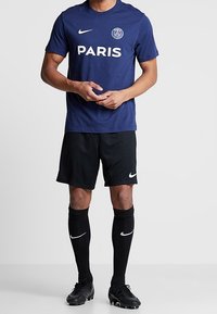 Námořnické modré tričko Nike s nápisem „PARIS“ v bílém písmu, doplněné o černé šortky, ponožky a kopačky s bílými „swoosh“.