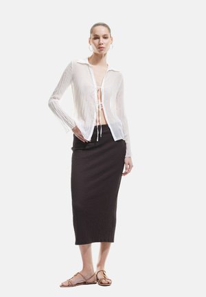 Transparante witte blouse met lange mouwen en een knoopsluiting aan de voorkant, gecombineerd met een textuurrijke donkerbruine midirok en sandalen met bandjes.