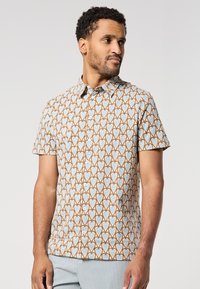 Homme portant une chemise à manches courtes avec un motif de cœurs bleus et bruns, associée à un pantalon gris clair, sur fond uni.