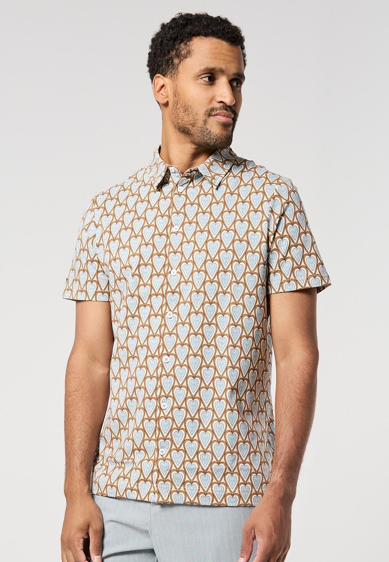 Hombre con una camisa de botones de manga corta con un patrón de corazones en azul y marrón, combinada con pantalones gris claro, frente a un fondo sencillo.