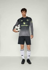 Camisola de futebol cinzenta de manga comprida com detalhes pretos, logótipo e texto verde. Calções pretos, meias brancas com riscas pretas e chuteiras pretas. A segurar uma bola de futebol.