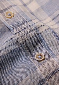 Camicia button-up in un motivi a quadri blu chiaro e bianco, realizzata in tessuto testurizzato. Presenta un colletto arrotondato e bottoni rotondi con cuciture dettagliate.