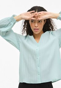 Gaiši zila plīsēta blūze ar V veida izgriezumu, pogu detaļām un elastīgām aprocēm. Audums ir gluds, nodrošinot vieglu tekstūru.