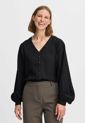 Black, textured blouse with a V-neck and long, puffed sleeves. Features front buttons and is paired with light brown trousers.
